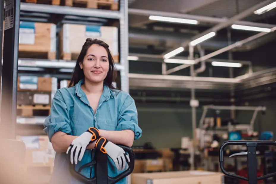 A woman stands confidently with her arms crossed, wearing a blue shirt and gloves, in a warehouse setting. She is smiling, surrounded by stacked boxes and various items in the background, which suggests a busy work environment.