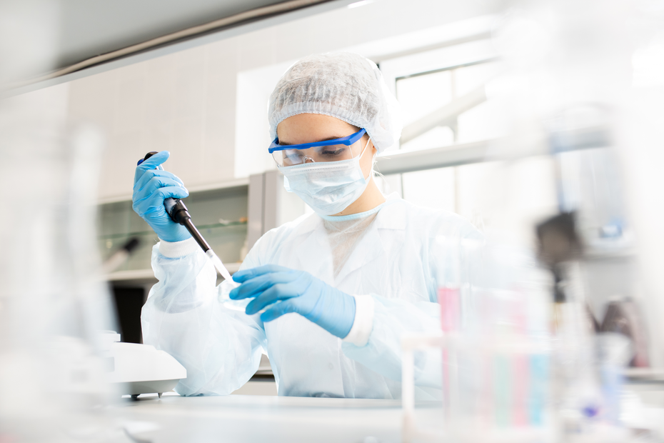 A person in a lab coat, gloves, goggles, and a mask is using a pipette to conduct an experiment in a laboratory setting. The background features lab equipment and glassware, emphasizing a scientific environment.