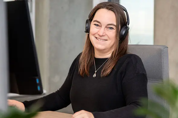 A woman with long brown hair is sitting at a desk, wearing a black sweater and headphones. She smiles while looking at the camera, with a computer monitor and some greenery visible in the background.