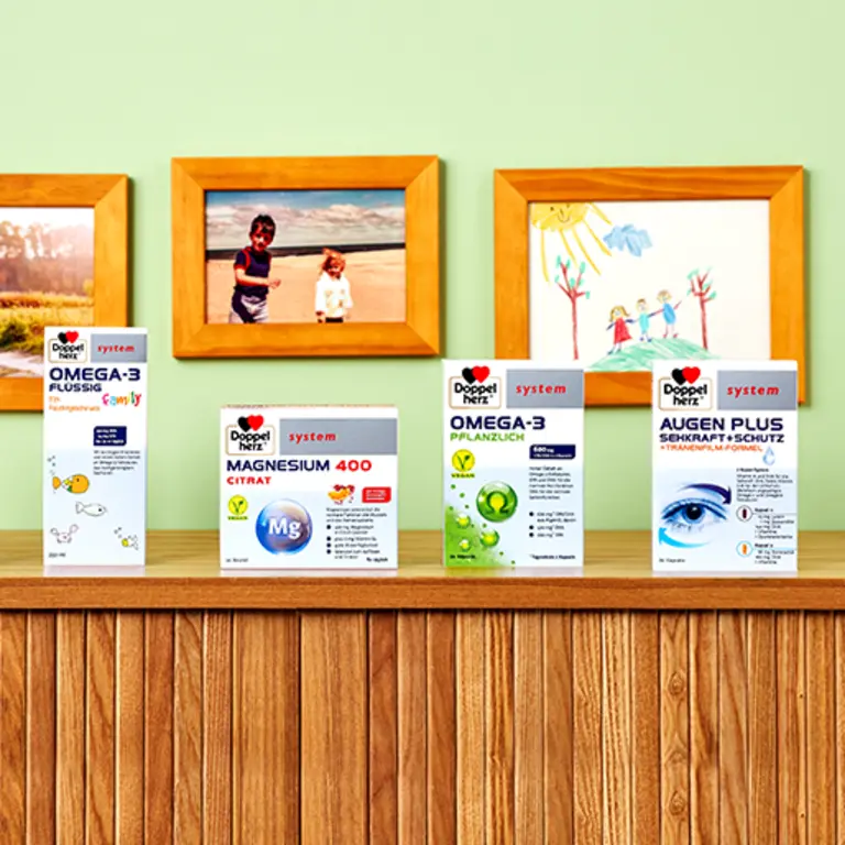 A wooden table holds four boxes of health supplements labeled "Omega-3 Flüssig," "Magnesium 400 Citrate," "Omega-3 Pflanzlich," and "Augen Plus," against a green wall. In the background, there are children's drawings and photographs featuring kids outdoors.