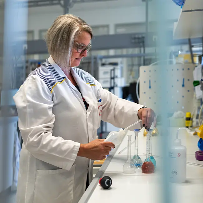 A scientist in a laboratory, wearing a white lab coat and glasses, is using a pipette to transfer liquid from a flask. Various glassware and lab equipment are visible on the workbench in the background.