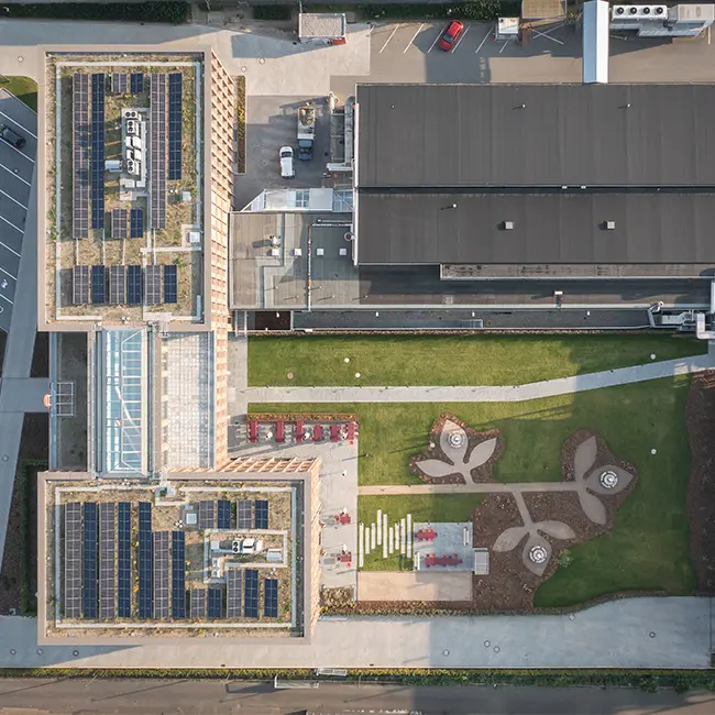 Aerial view of a modern building complex featuring two structures with green roofs and solar panels. The landscaped area includes pathways, seating areas, and artistic designs in the ground. Nearby, a parking area is visible, emphasizing the eco-friendly design elements.