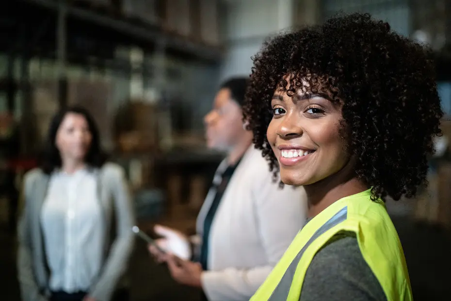 A smiling woman with curly hair and a yellow safety vest stands in a warehouse setting, while two other women are engaged in conversation in the background. The atmosphere appears professional and collaborative.
