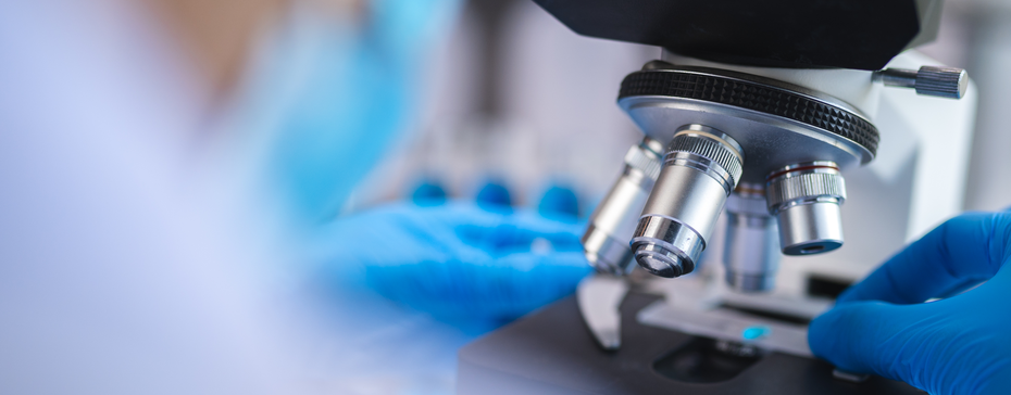A close-up of a laboratory microscope with a person's gloved hand adjusting the slide. The background is blurred, focusing on the microscope's lenses and the hand in a blue glove.