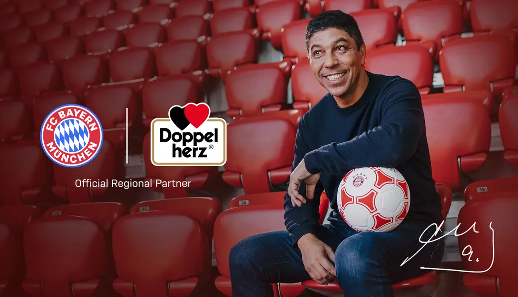 A smiling man sits in a stadium seat holding a soccer ball. There are logos for FC Bayern München and Doppelherz, indicating an official partnership. The background features red stadium seating, emphasizing a sports theme.