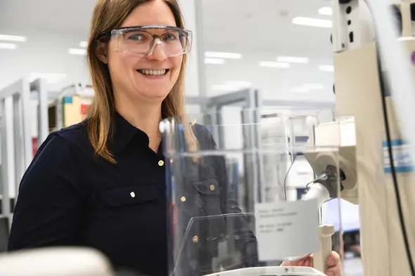 A smiling woman wearing safety glasses stands in a laboratory setting, next to a piece of lab equipment. She is dressed in a dark shirt and appears engaged in her work. The background shows shelves and equipment typical of a research environment.