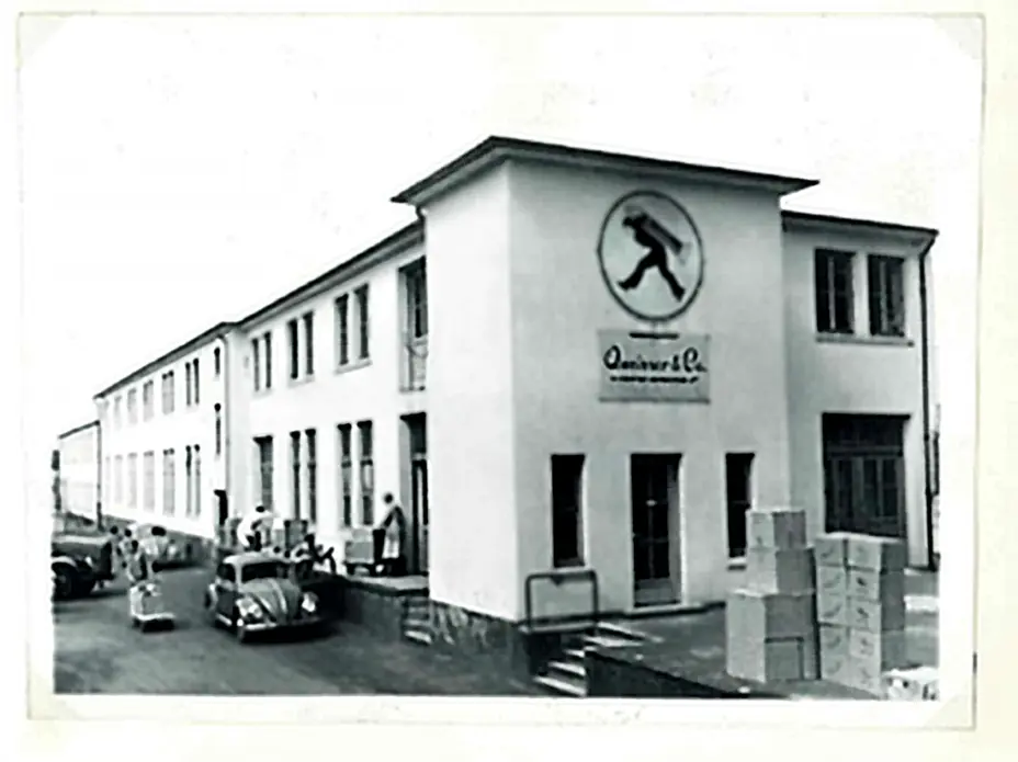 A black and white historic photograph of a building labeled "Quinivar & Co." featuring a logo of a person walking. The structure has multiple windows, a set of stairs, and boxes stacked nearby. Vintage cars are parked in front, with a few people visible outside.